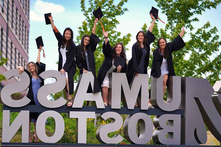 Students in graduation gowns stand and cheer at the UMass Boston sign.